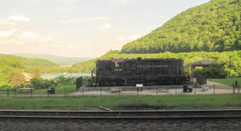 Beyond the freight engine, note a cut in the trees in the right background, the western end of tracks wraping around Horseshoe Curve west of Altoona, PA. Finished in 1854 by the then-Pennsylvania Railroad, this wrap around the mountains made the transportation of heavy trains possible for development of the Midwest and Western States. The Pennsylvanian slows down to look at this engineering achievement. Photo: Ralph Spielman