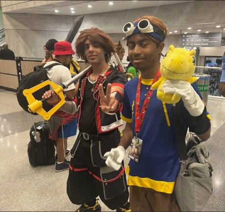 Two cosplayers pose together: one as Sora from “Kingdom Hearts” holding a keyblade (left) and the other in a blue and yellow outfit, dressed as Tai from Digimon carrying a yellow Digimon plush toy--his partner Agumon.