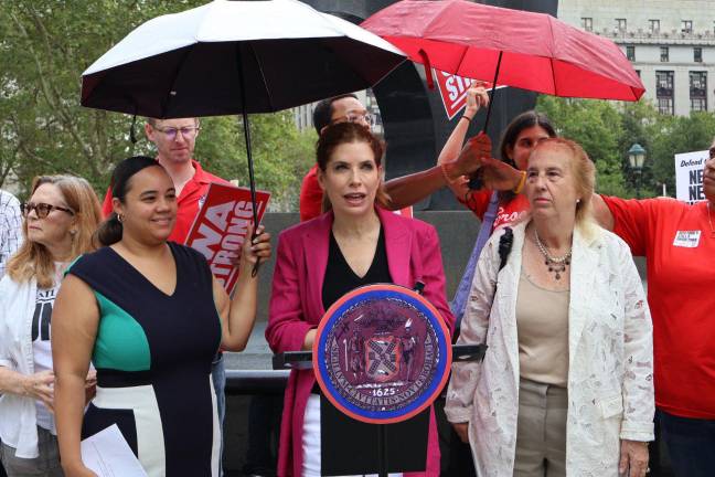 Julie Menin at the podium, Aug. 14, 2025. Flanking her, Council Members Carmen De La Rosa, left, and Gale Brewer. News Guild New York President Susan DeCarava holds unbrella right.
