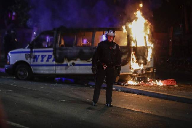A police officer watches a crowd as a police vehicle burns near Fort Greene Park in Brooklyn after protesters rallied outside Barclays Center over the death of George Floyd, who died Memorial Day while in police custody in Minneapolis, Friday, May 29, 2020.