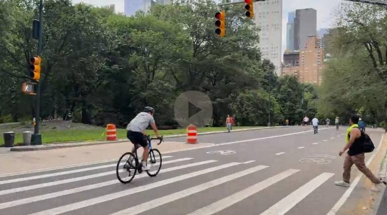 A biker in Central Park runs a red light visible in traffic signal above as a pedestrian who has the right of way (white walk light visible on left) steps into cross walk.